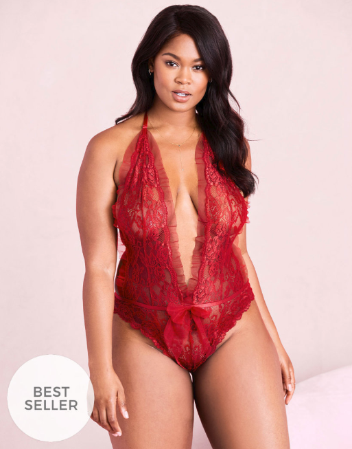 Adore Me - alternative to Victoria's Secret for plus size lingerie. A plus-size model with brown skin and dark brown hair poses in front of a pale pink background, wearing a red lace teddy.