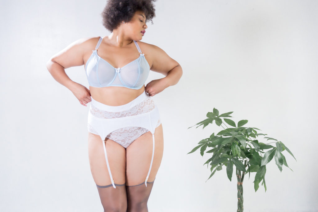 Plus size lingerie company Impish Lee. A woman with brown skin and bushy hair stands in front of a white wall next to a miniature tree. She is wearing a light blue bra, white lace panties, a white lace garter belt, and stockings.