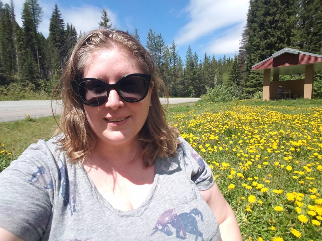 A plus-size woman in a gray t-shirt and big sunglasses stands in a field of dandelion flowers in front of a picnic shelter and paved road.