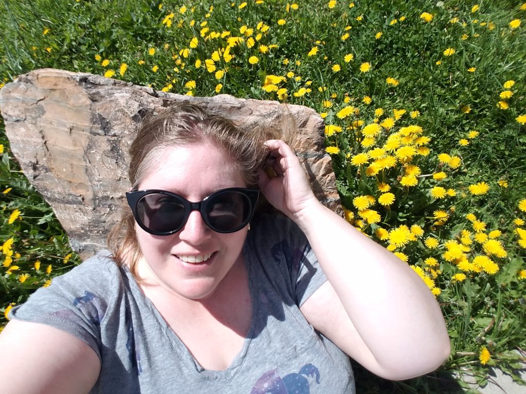 A fat woman with blonde hair, sunglasses and a gray shirt leans against a rock in a field full of dandelions.
