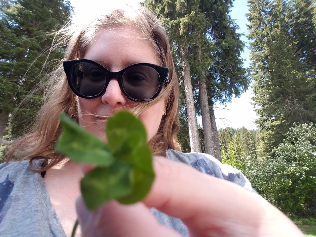 The same woman stands in front of tall pine trees and holds a four-leaf clover up to the camera, which is out of focus, darn it.