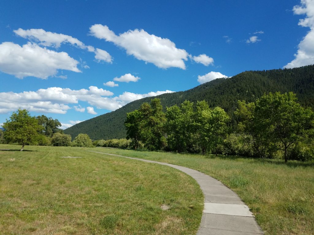 A random Montana parking area. Looks like maybe it used to be a full rest area, given the concrete sidewalks. The sidewalks are surrounded by green grass in front of trees and a mountain ridge.