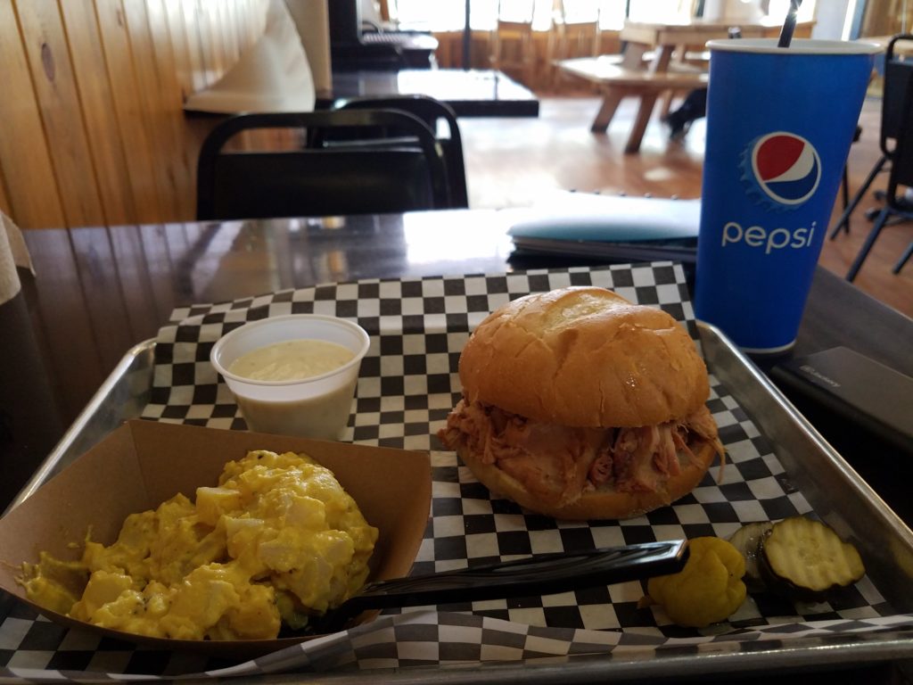 The predominant flavor of potato salad really shouldn't be black pepper, but it was a good try. Yellow potato salad, a barbecue sandwich, and a Pepsi cup are shown on a table in a restaurant with wood-paneled walls.