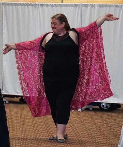 A fat white woman with shoulder-length brown hair throws her arms wide in joy and confidence as she walks in a fashion show in front of a white sheet. She's wearing a black sleeveless top and pants with a bright pink lace kimono-style jacket.