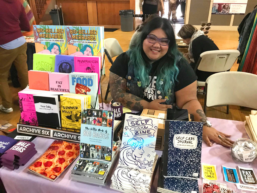 A smiling woman with brown skin, purple glasses and black and blue hair is sitting behind a table at an event with a variety of zines and stickers.