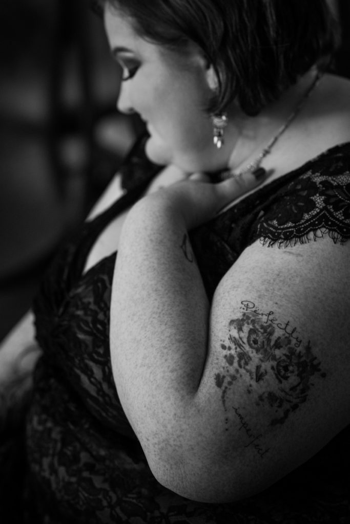 A fat white woman wearing a black lace top, necklace and earrings, with eye makeup, and short dark hair, sits with one hand pressed to her upper chest. An owl watercolor tattoo on her upper arm is accompanied by the words "perfectly imperfect."