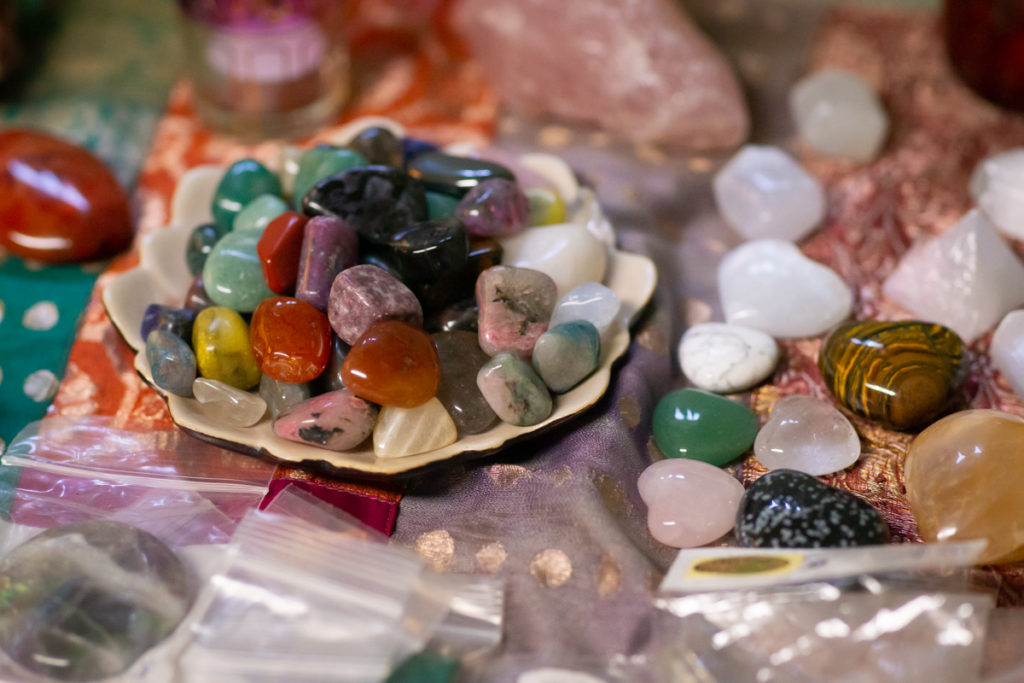 A number of colorful crystals for crystal healing and energy work rest on a colorful fabric cloth in green, red and orange tones. The stones include tiger's eyes, obsidian, aventurine and quartz hearts and tumbled pink, yellow, red, orange, purple, white, clear and green stones.