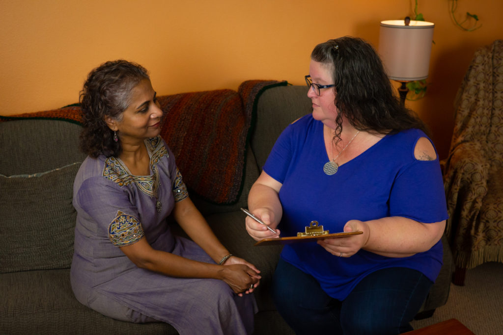 A woman of Indian descent in a purple embroidered dress and a fat white woman in jeans and a dark blue top sit on a brown couch in a waiting room and discuss an appointment. The white woman holds a clipboard and pen.