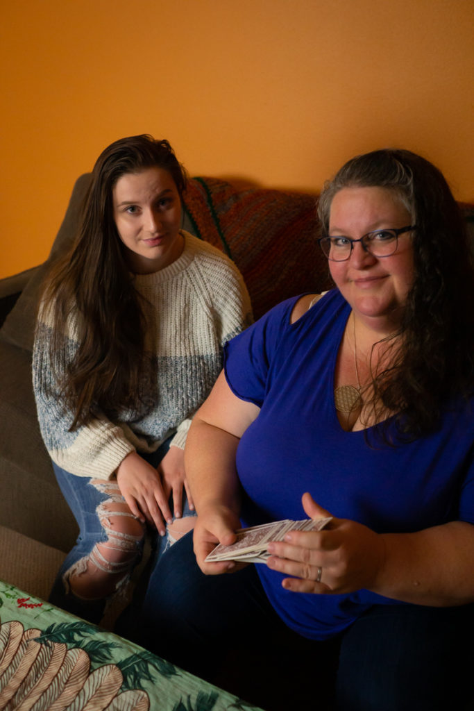 Two plus-size white women sit on a brown couch in front of an orange wall and get ready to interact with tarot cards, both looking at the camera. The younger of the two women has on a sweater and jeans. The older of the two women has on a dark blue top and jeans, and is shuffling a deck of cards.