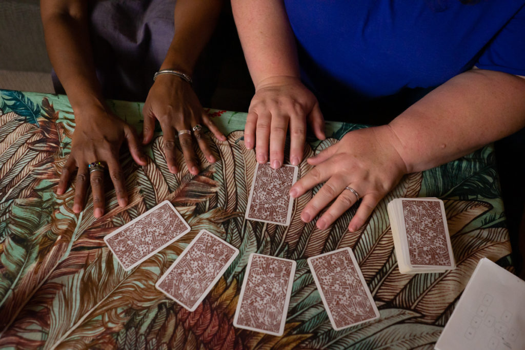 An Indian-American woman and fat white woman's hands are shown resting on a coffee table covered in a wing-pattern scarf in green, orange and brown tones. A set of tarot cards lies face-down on the table.