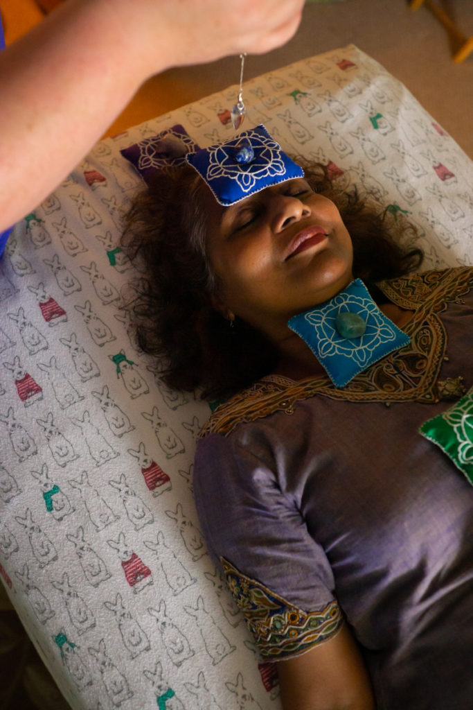 An Indian-American woman in an embroidered purple dress lies on a table covered in a cartoon rabbit-printed fabric. Chakra beanbags and gemstones lie on her forehead and chest. A white woman dangles a crystal near her face.