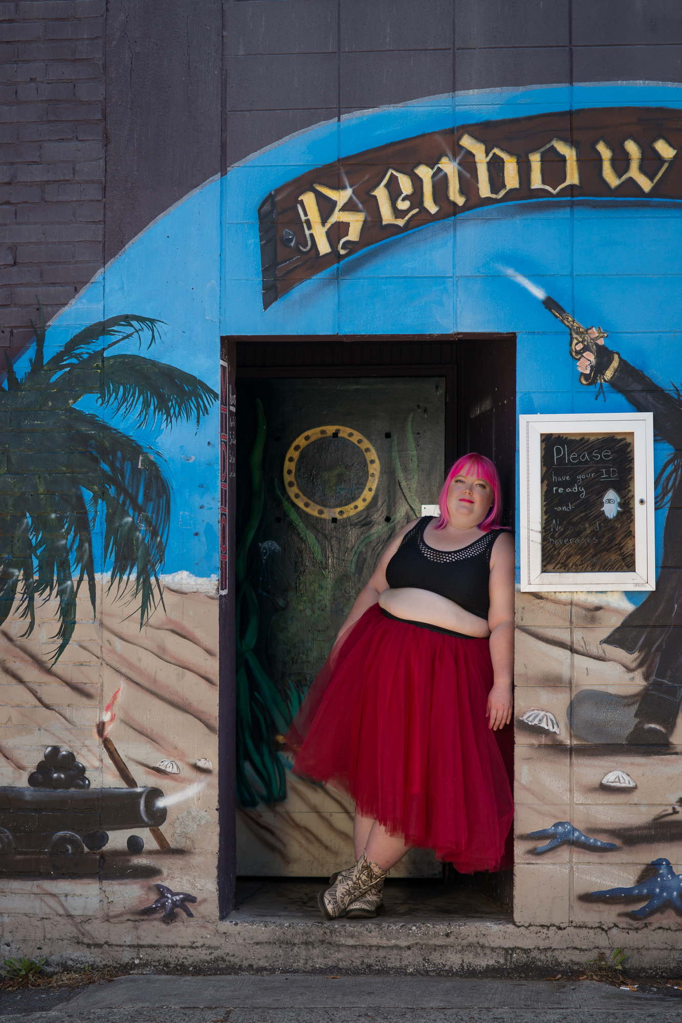 A fat woman with pink hair leans against a doorframe in a red skirt and a crop top