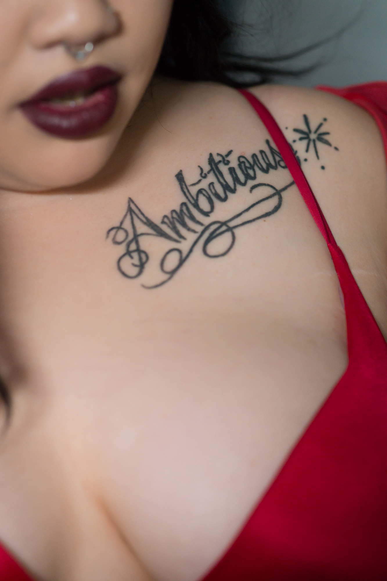 A Black woman wearing dark lipstick poses in red lingerie, with a tattoo on her shoulder that reads "Ambitious"