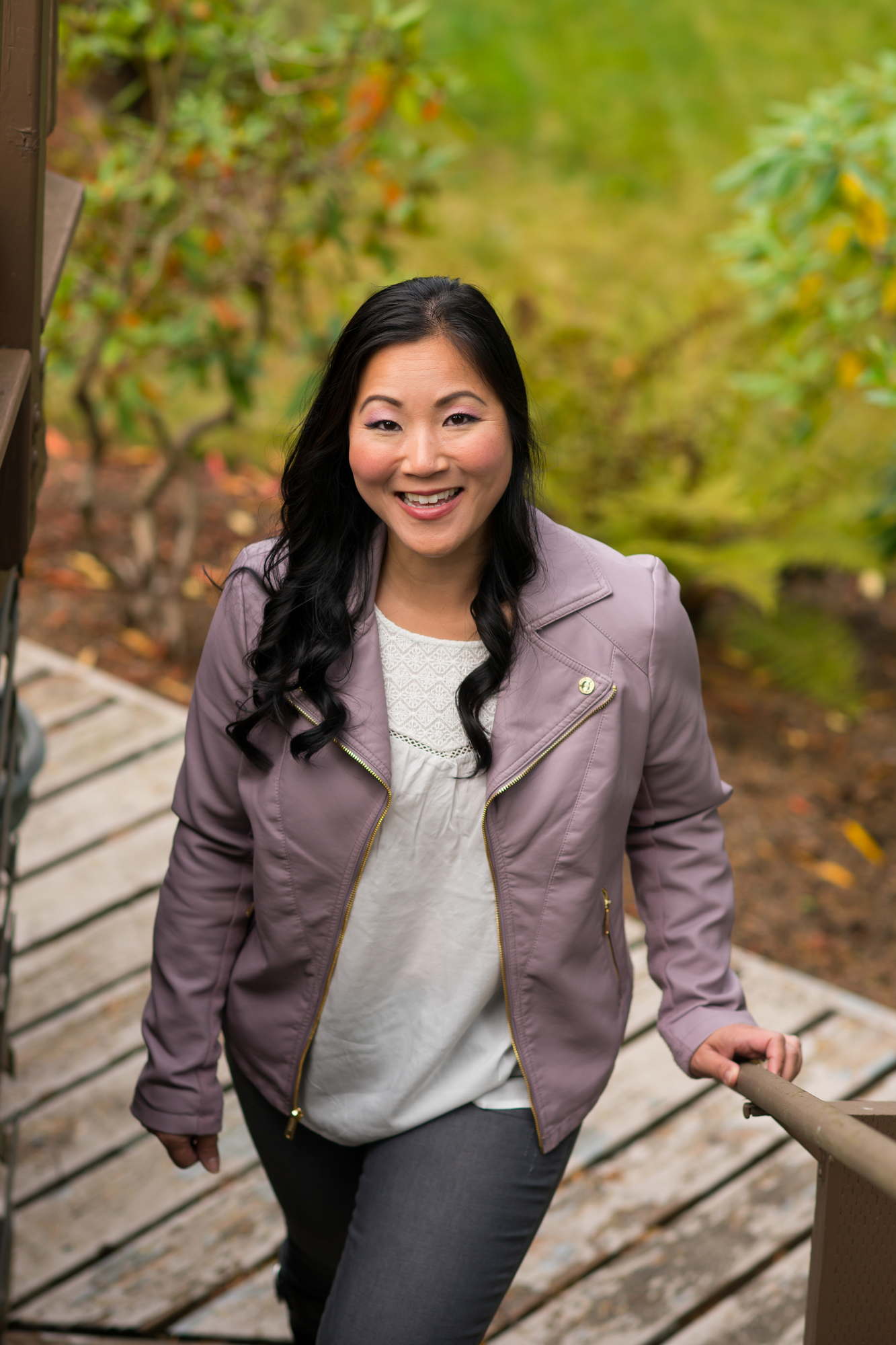 A Brown woman wearing a lavendar jacket poses on a deck outdoors in the PNW