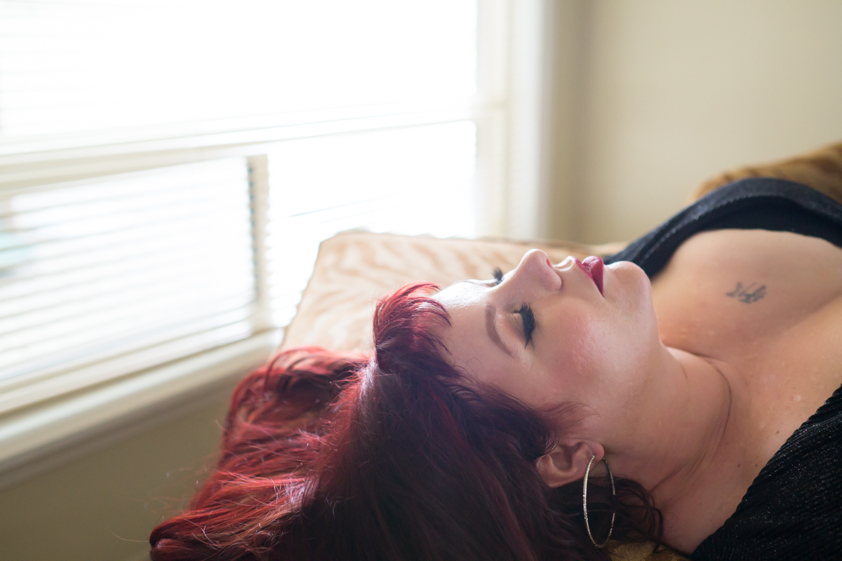 A woman with burgundy hair lays with her eyes shut framed by a window during a fat positive photo session in Seattle