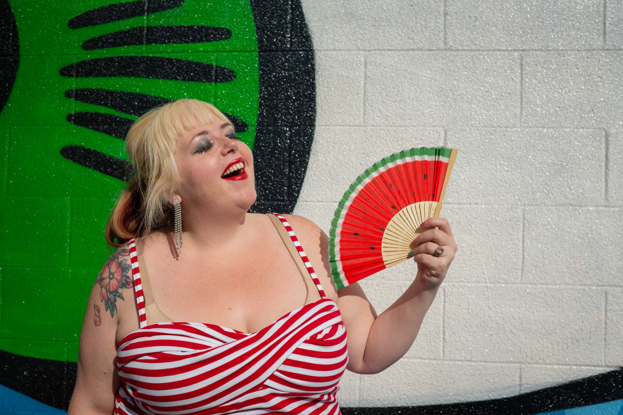 A fat woman wearing red lipstick and a red striped blouse laughs while holding a fan 