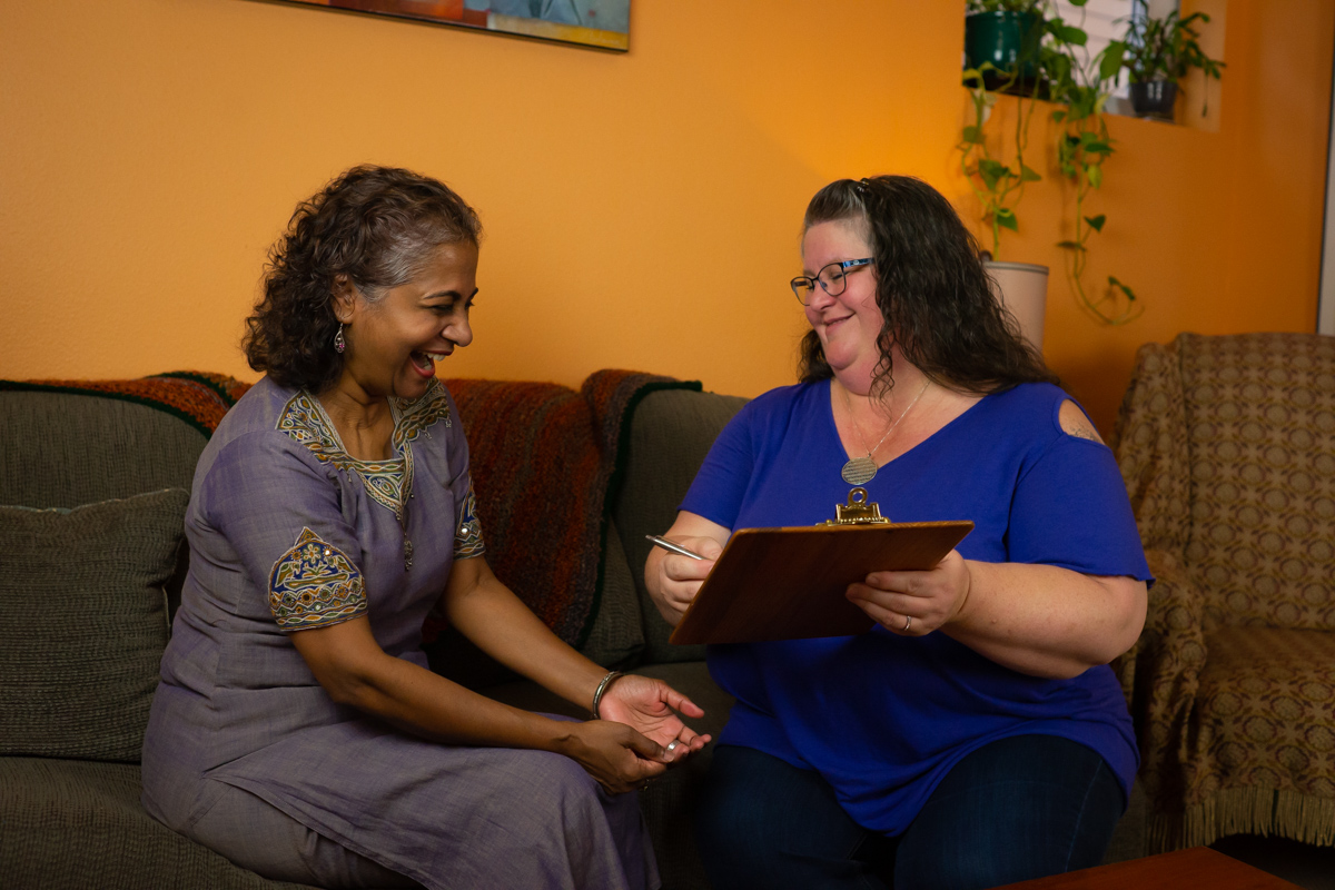 A fat woman holding a clipboard smiles at a laughing Black woman in an office
