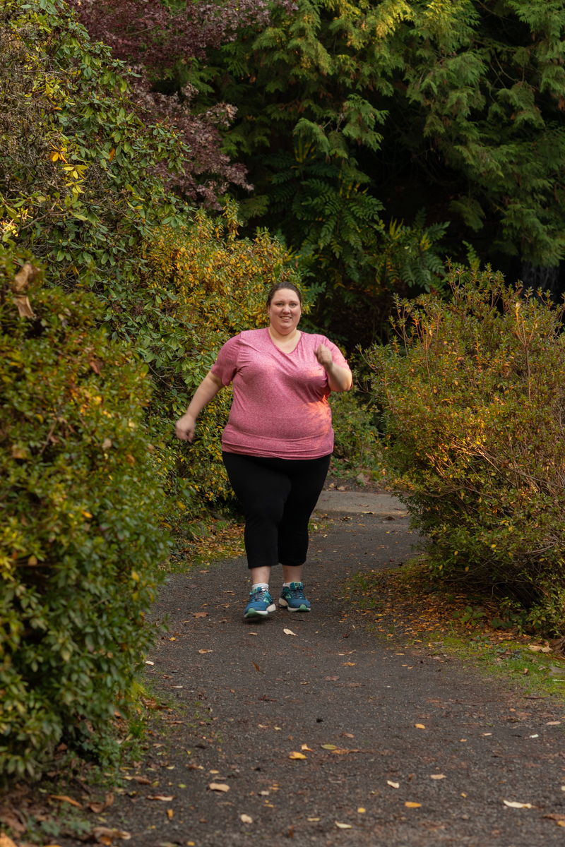 A fat woman in a pink workout top hikes in the woods in the PNW