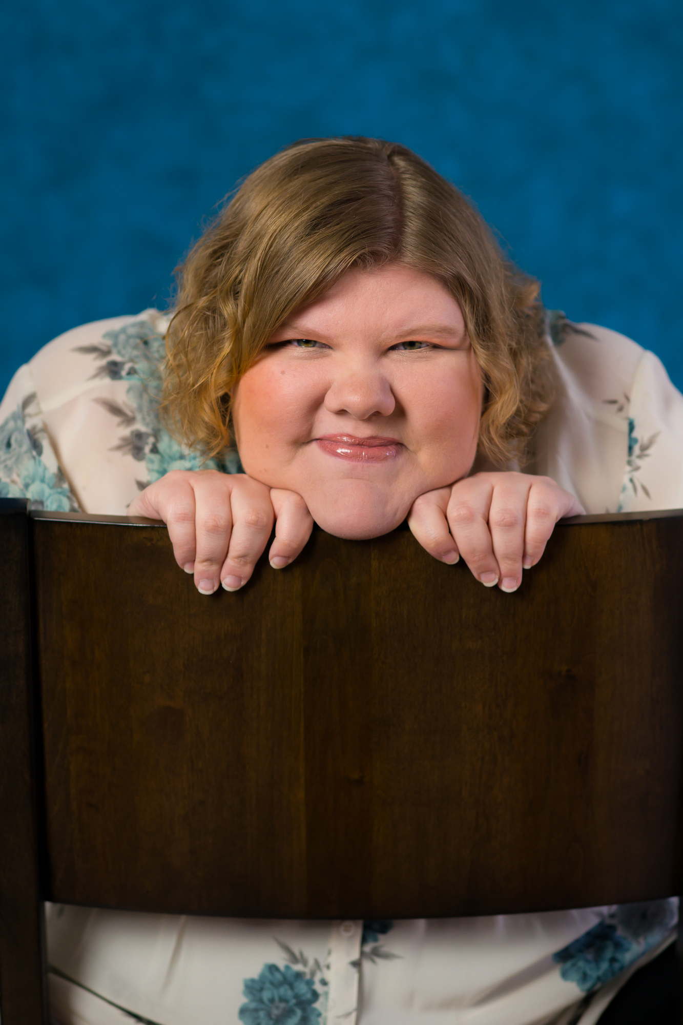A fat woman looks at the camera playfully while posing over the back of a chair