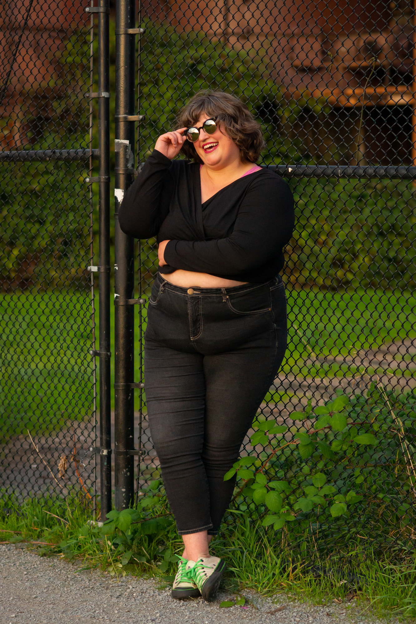A fat woman wearing sunglasses leans against a fence during a portrait photoshoot outdoors