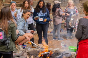 A group of girls gather around a fire outdoors in the PNW