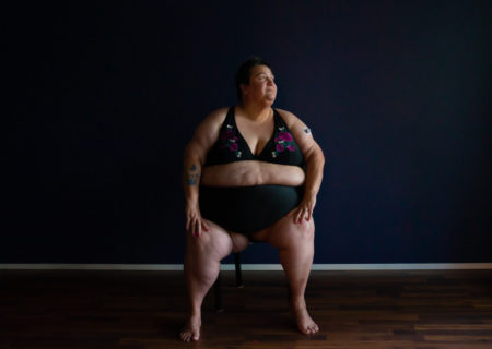 A fat woman with pale skin sits on a chair in a dark room, body facing forward and face turned to the right. She is wearing a black bikini with a floral design.