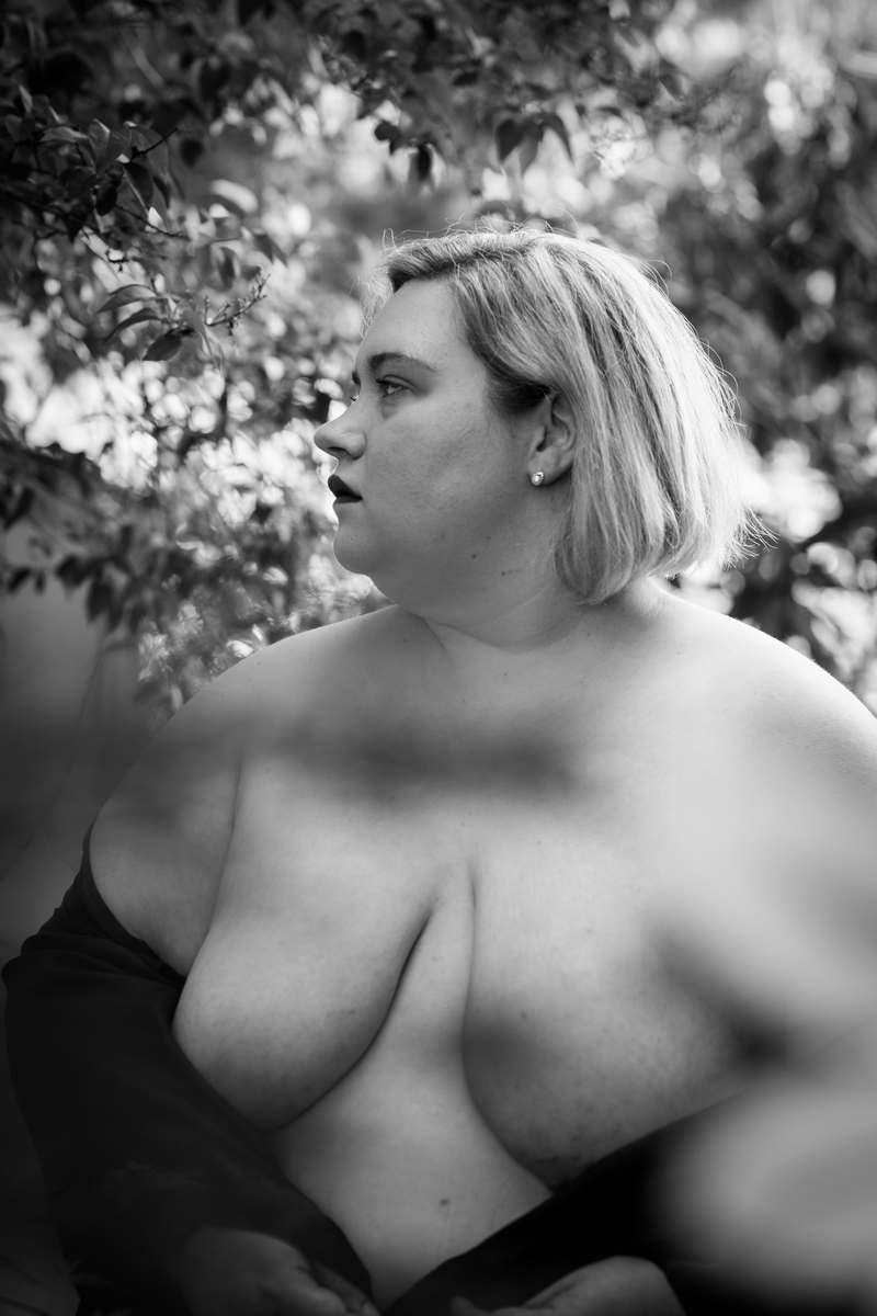 Image description: A fat white women with shoulder-length blonde hair is shown in a black and white image with a black drape just covering the tips of her breasts and part of her torso. She is looking off to one side with a solemn expression, with tree limbs behind her.