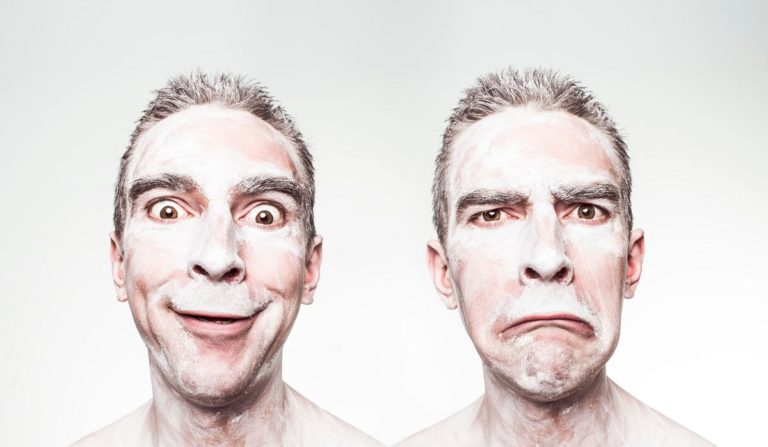 A thin white man with close-cropped hair is shown from the neck up in two shots, side by side. In the first, he has a manic smile. In the second, he has an exaggerated frown. In both shots, his face and neck are covered with a pasty white substance.