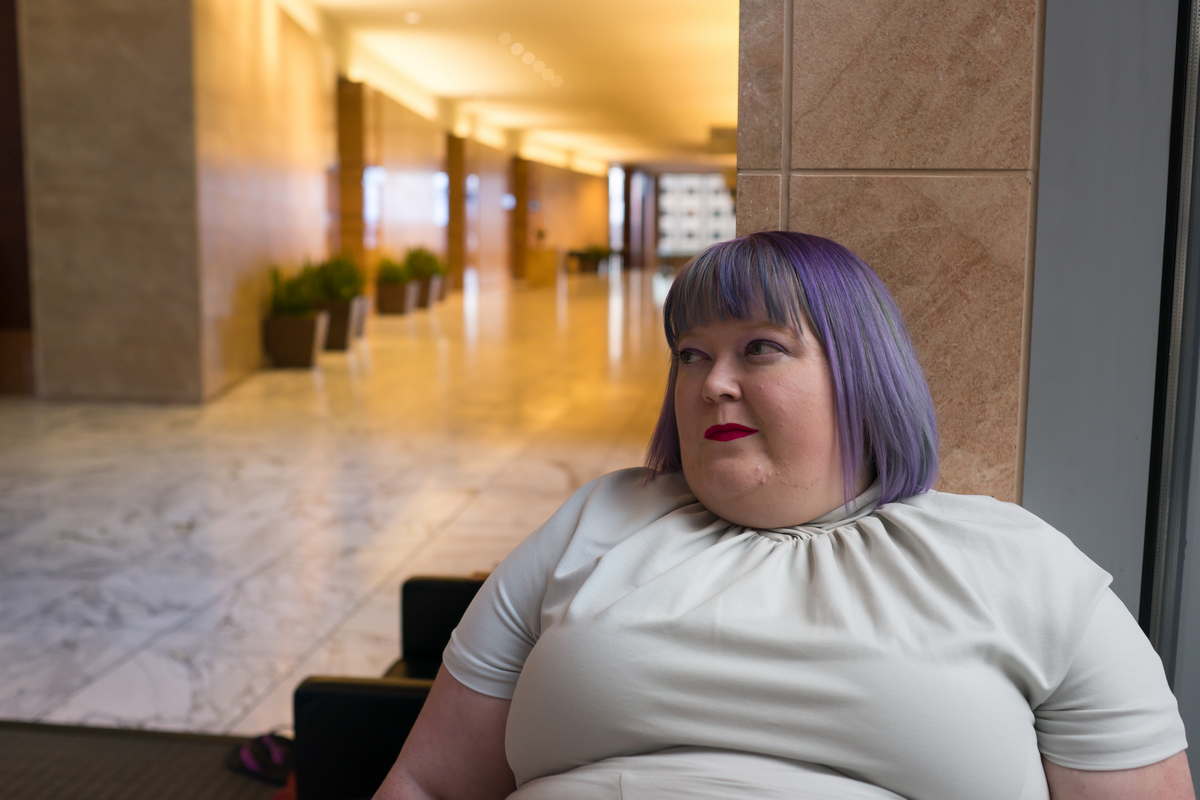 Image description: A fat white woman with purple hair, red lipstick and a gray short-sleeved dress is shown from the bust up. She is sitting in the lobby of a large corporate office building. An empty hallway with marble floors and large plants in planters stretches out behind her. End image description.