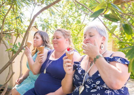 Three fat women sit outside under trees in summer dresses and eat ice cream cones.