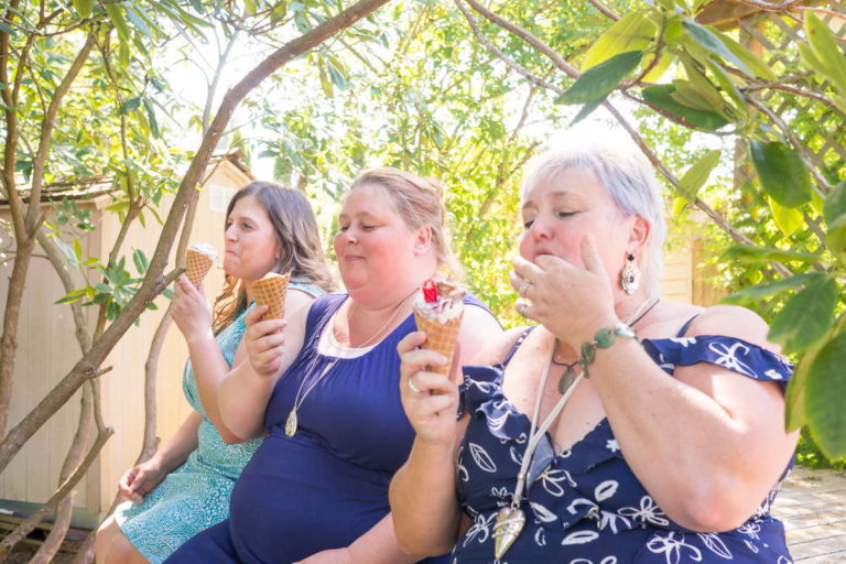 Three fat women sit outside under trees in summer dresses and eat ice cream cones.