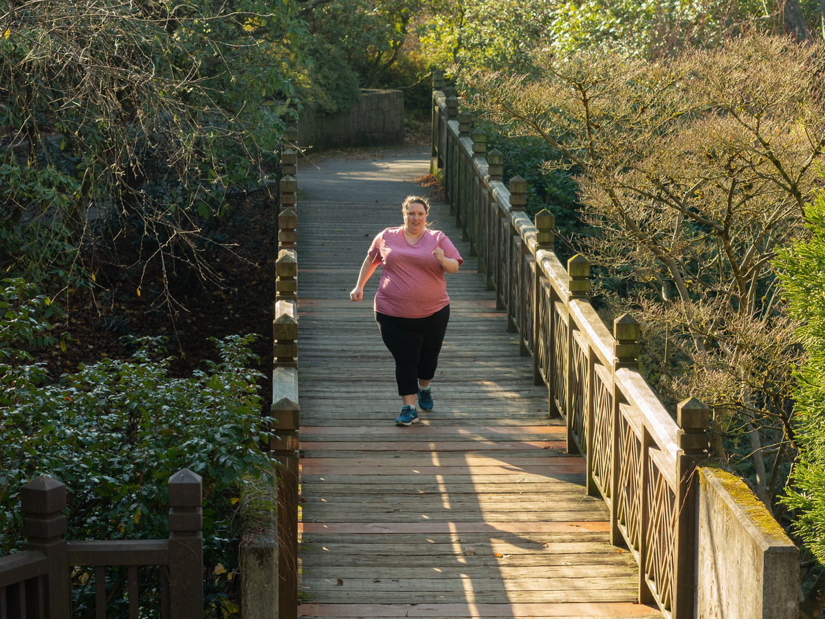 A fat white woman in pink t-shirt and black pants runs towards the camera along a wooden bridge surrounded by a green forest.