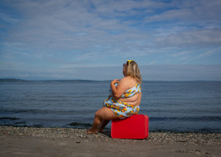 A fat blonde woman in a bikini with visible side rolls sits on a vintage red suitcase at the edge of the water on a beach, looking away.