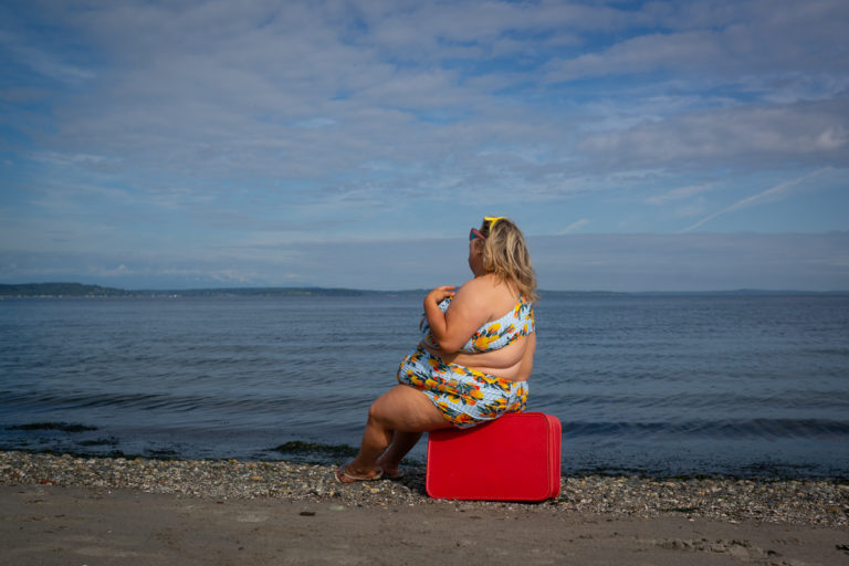 A fat blonde woman in a bikini with visible side rolls sits on a vintage red suitcase at the edge of the water on a beach, looking away.