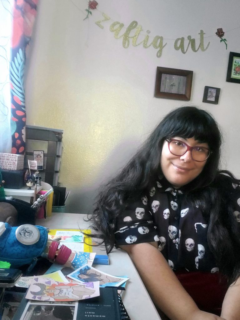 Picture of Kristine sitting at her desk with a piece of artwork in front of her. She has long dark hair with bangs and is wearing red glasses and softly smiling at the camera. Above her are photos on the wall and a sign reading "zaftig art".