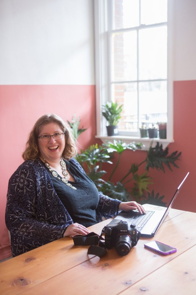 Lindley, a small business owner who's experienced burnout and recovered, sits at a cafe table with her phone, camera and laptop.