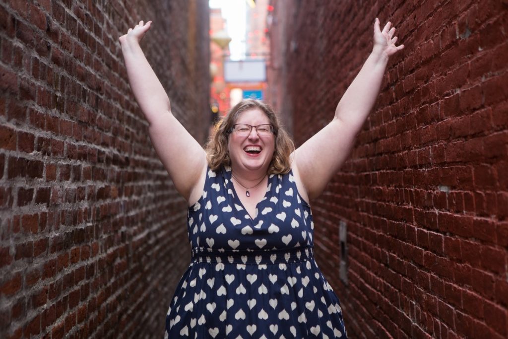 Lindley, a fat white female entrepreneur, stands in a narrow brick alley in a heart-patterned dress with her arms lifted.