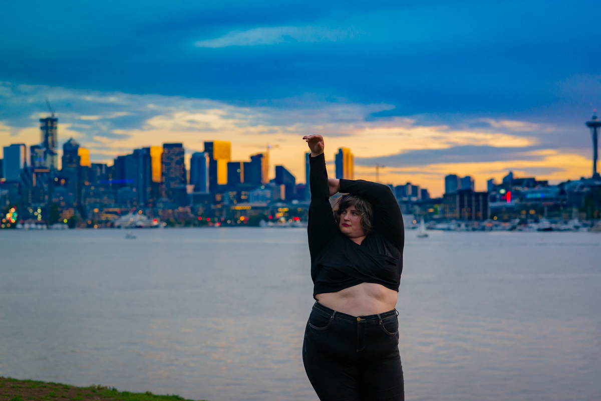 A plus-size woman poses in front of a sunset-lit city skyline and water with one arm in the air and the other wrapped around it. She’s wearing blue jeans and a black crop top sweater.