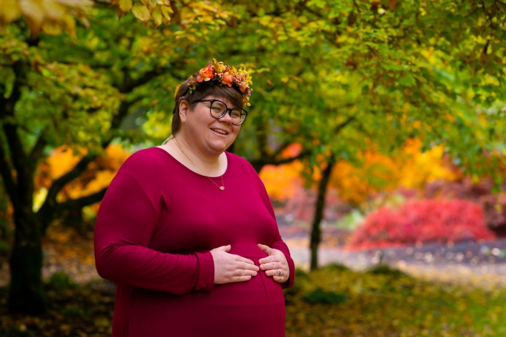 A plus-size woman with pale skin, short brown hair, and glasses stands in the middle of fallen yellow leaves with autumn trees and bushes around her. She is wearing a necklace, a long pink dress, sandals, and a flower crown, and is cradling her belly with her hands. She's looking away from the camera and smiling.