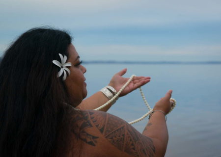A fat Hawaiian woman with brown skin and black hair lifts a long pearl necklace toward a cloudy sky and blue water.