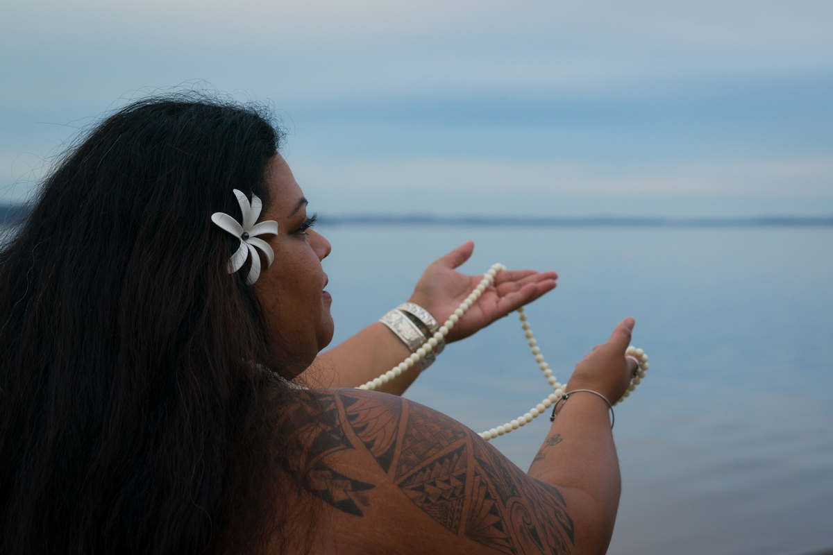 A fat Hawaiian woman with brown skin and black hair lifts a long pearl necklace toward a cloudy sky and blue water.