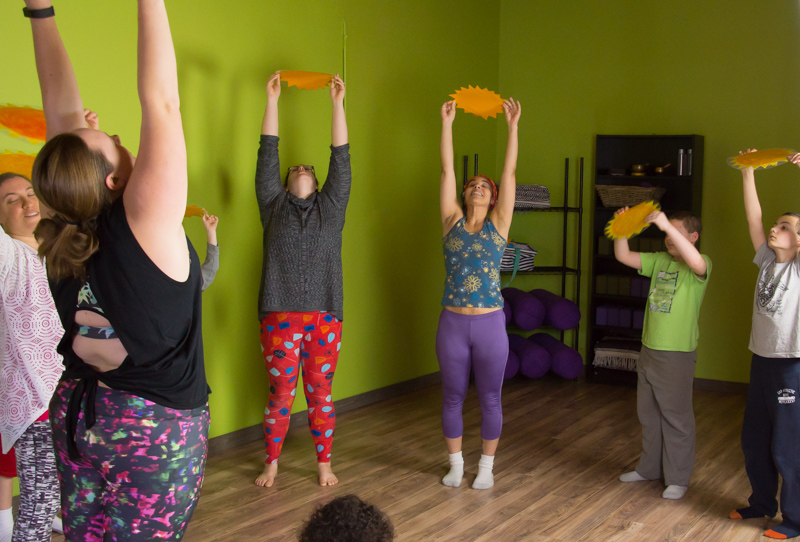 People in a family yoga class stretch their arms high, holding paper suns.