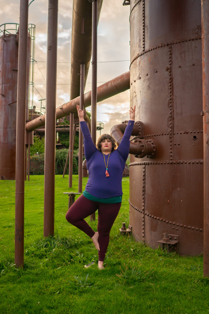 A fat woman does a tree yoga pose among large industrial pipes.