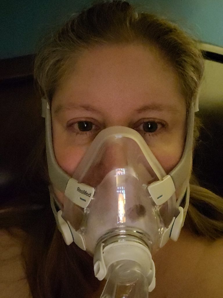 Image description: Lindley without glasses in a full face mask. covering mouth and nose, that attaches to a CPAP machine.