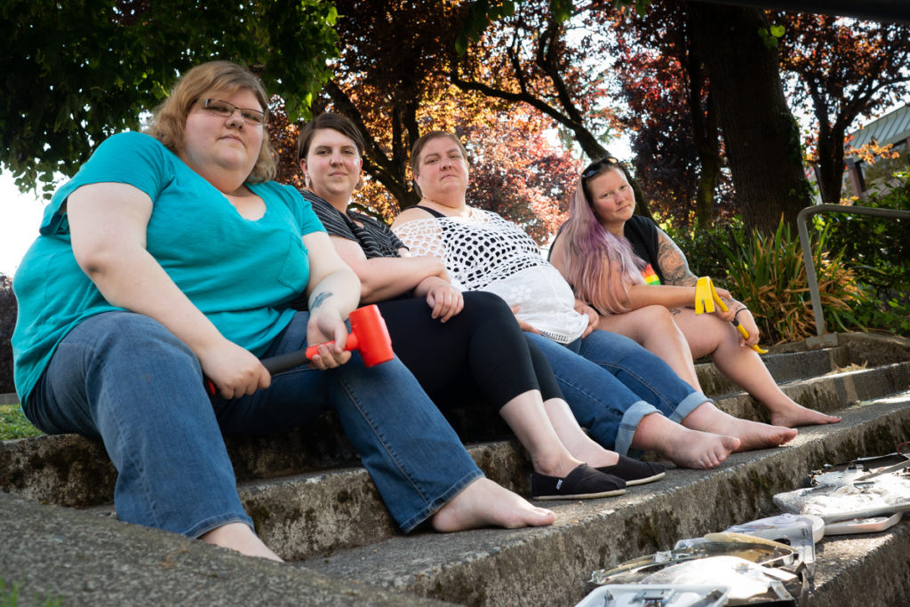 A group of people in larger bodies sit on concrete steps with hammers and smashed bathroom scales.