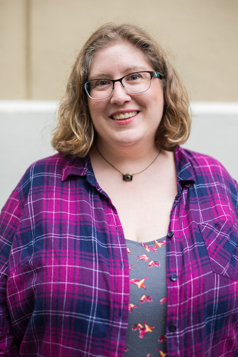 Lindley, a fat white woman, looking into the camera and smiling. She's wearing glasses, a pink and blue top and a camera necklace.