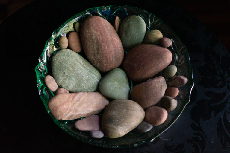 A green glass bowl of smooth pebbles in soft red and green tones and mixed sizes, stones Lindley found in a river in Idaho