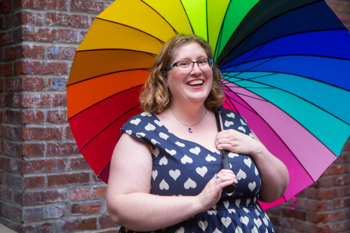 Image description: Lindley, a fat white woman, stands in front of a brick wall in a heart polka-dot dress. She's holding a rainbow umbrella and smiling.