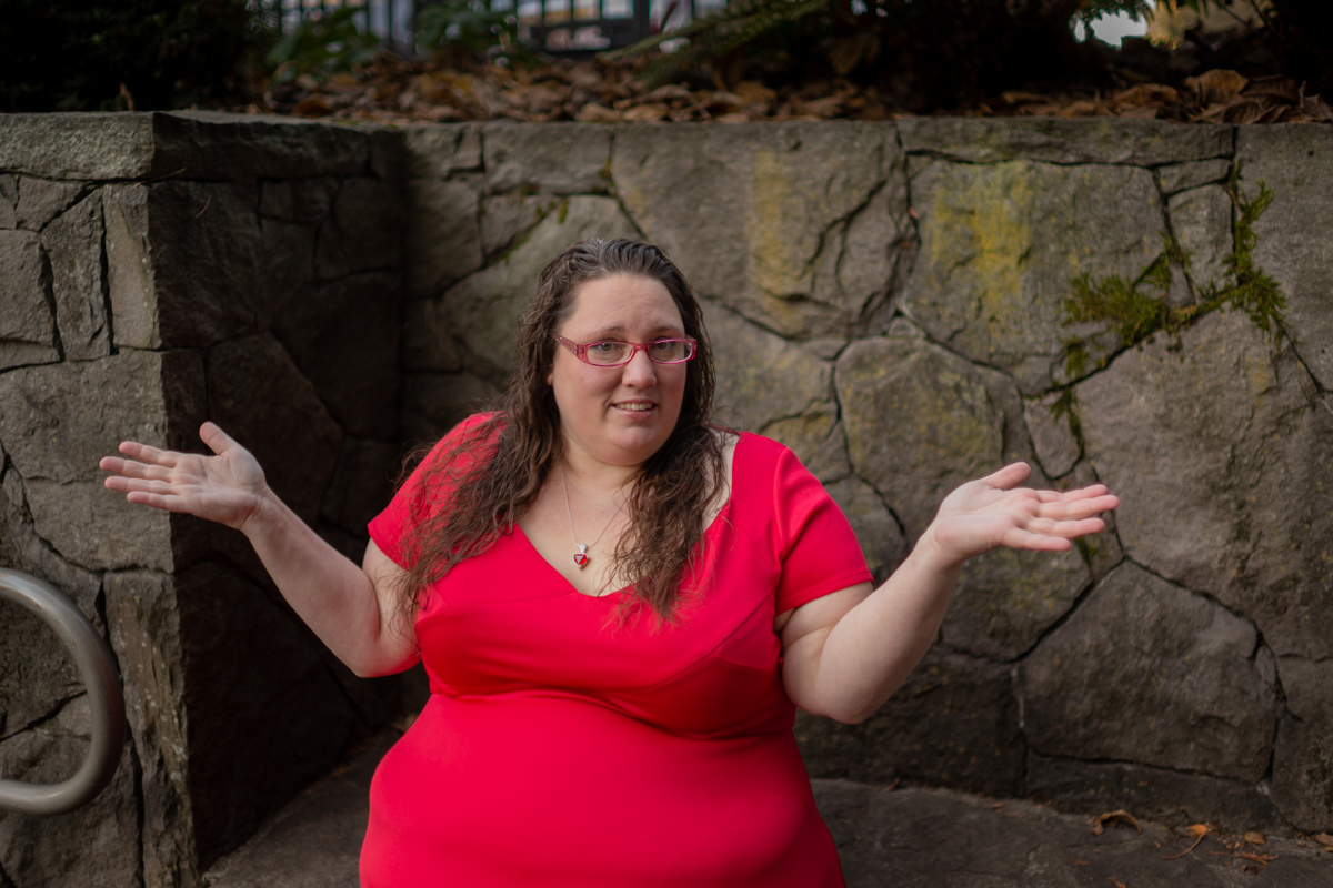 A fat white woman in a short-sleeved red dress sits in front of a stone wall and shrugs.