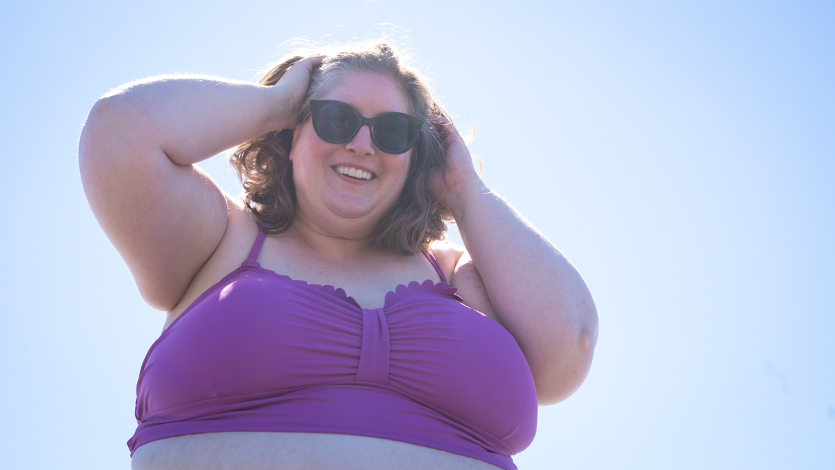 Lindley, a fat white woman with sunglasses and a purple swimsuit top, is shown from below holding her hair up and smiling at the camera.